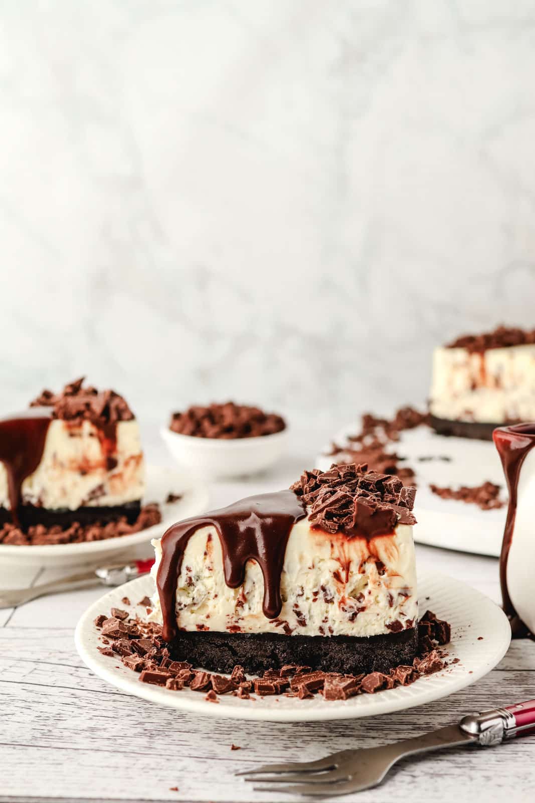 Choc chip cheesecake with chocolate sauce drizzled over the top, jug of chocolate fudge sauce on the side and rest of cheesecake in background.