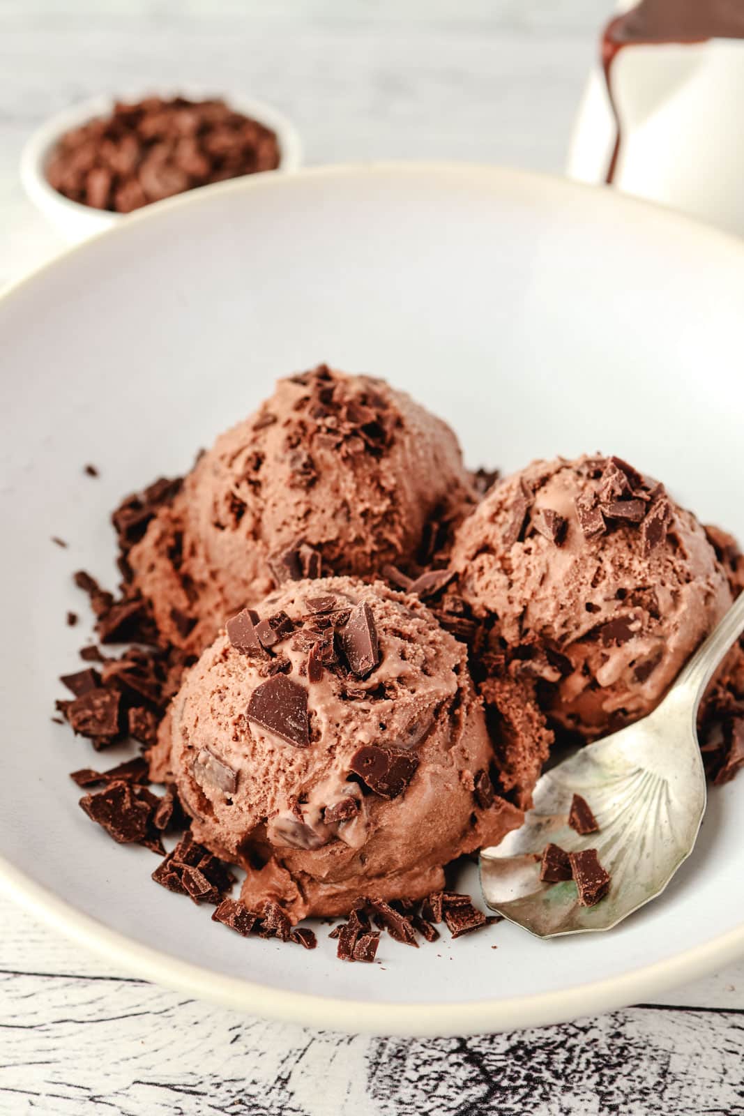 Chocolate Chips For Ice Cream 1 Three scoops of double chocolate ice cream in a bowl made with homemade chocolate chips for ice cream, with a spoon on the side and extra chips and chocolate sauce in the background.