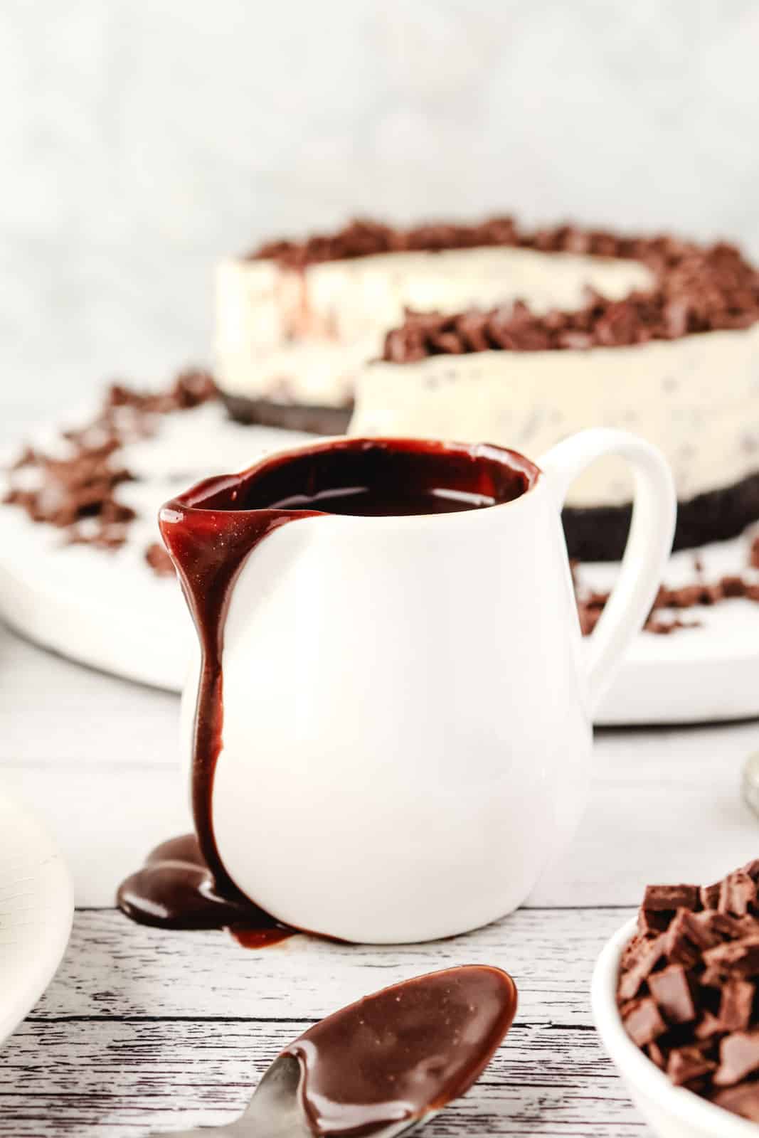 Homemade Chocolate Sauce 2 Jug of chocolate sauce with chocolate drip on the side, chocolate-dipped spoon in the foreground and chocolate chip cheesecake in the background.