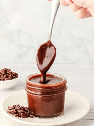 Chocolate sauce dripping from a spoon into a small mason jar on a plate with chocolate chunks.
