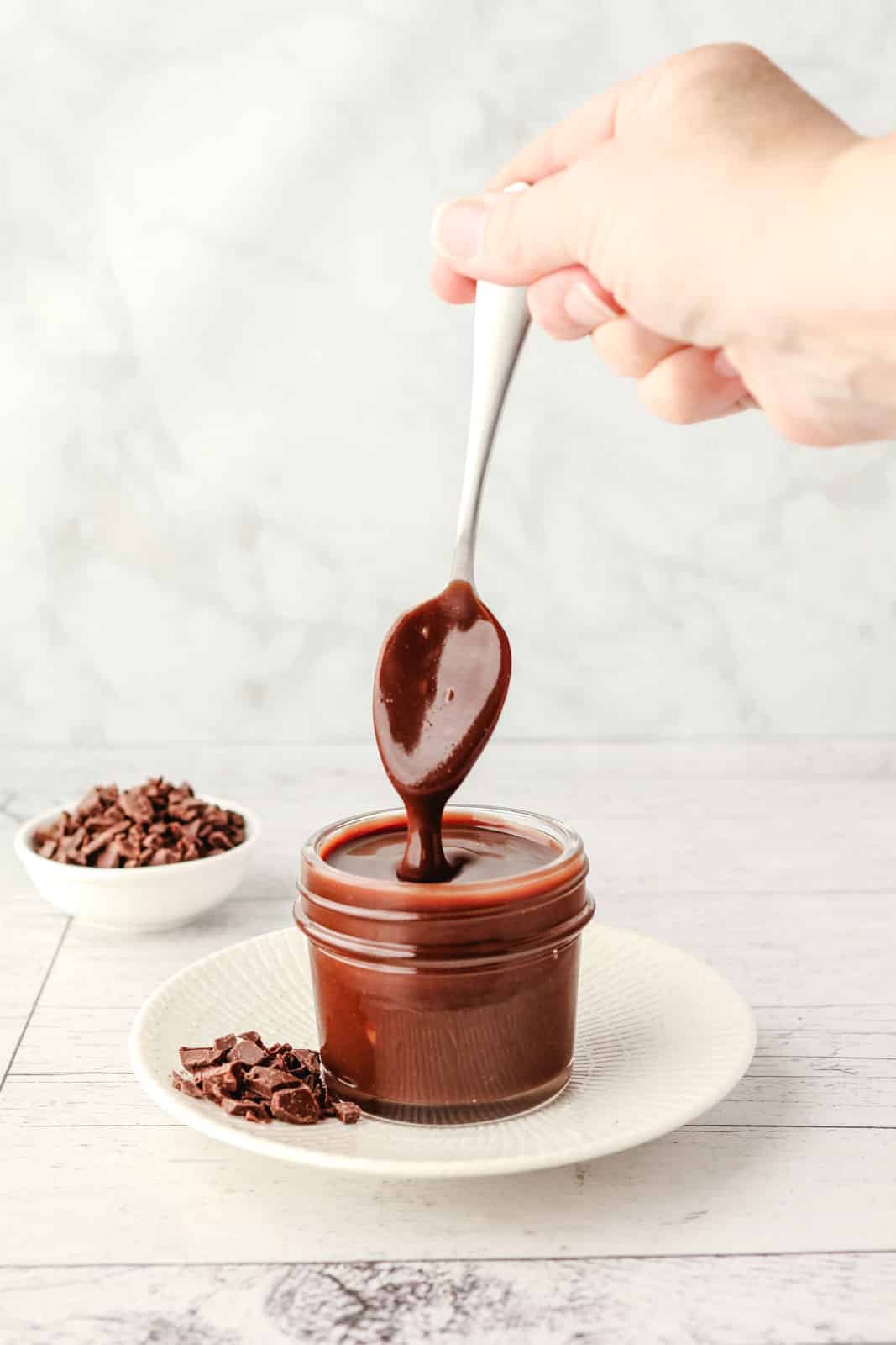 Homemade Chocolate Sauce 6 Chocolate sauce dripping from a spoon into a small mason jar on a plate with chocolate chunks.